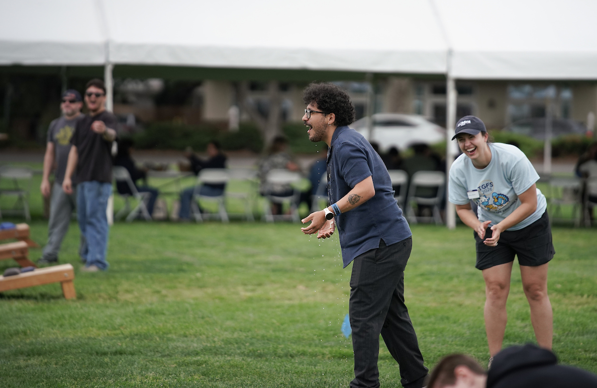 Two people playing cornhole on a grassy lawn, one mid-throw, tents and spectators behind