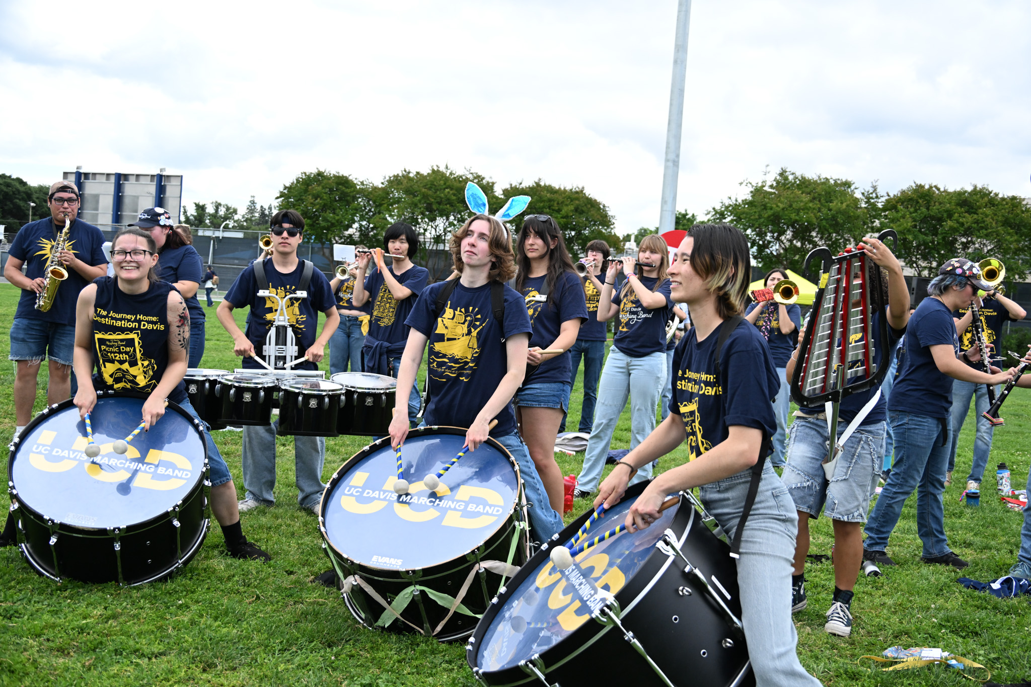 Marching-band drummers smiling while playing bass and snare drums on a sunny grassy field