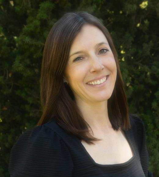 Smiling woman headshot, shoulder-length brown hair, black top, leafy green background